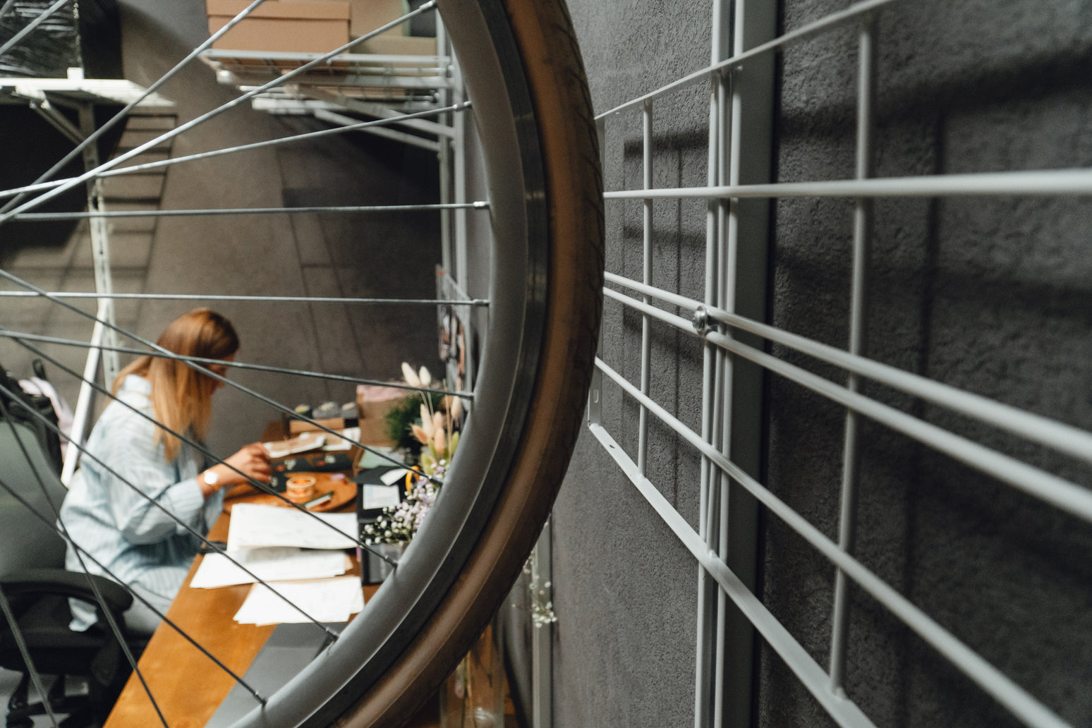 Person working at a desk through a window with a bicycle wheel in the foreground.