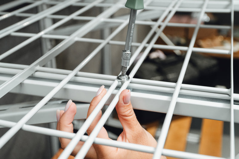 A close-up of a person's hand using a screwdriver to secure a screw onto a white metal wire shelving unit.